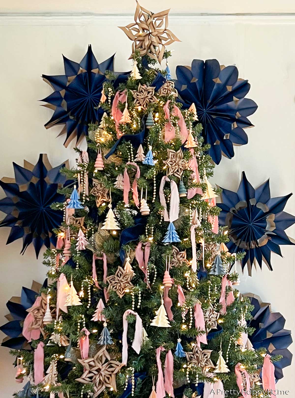 pink and navy christmas tree with handmade origami ornaments, paper snowflakes, and mini wood trees set against a backdrop of giant snowflakes made from blue gift bags