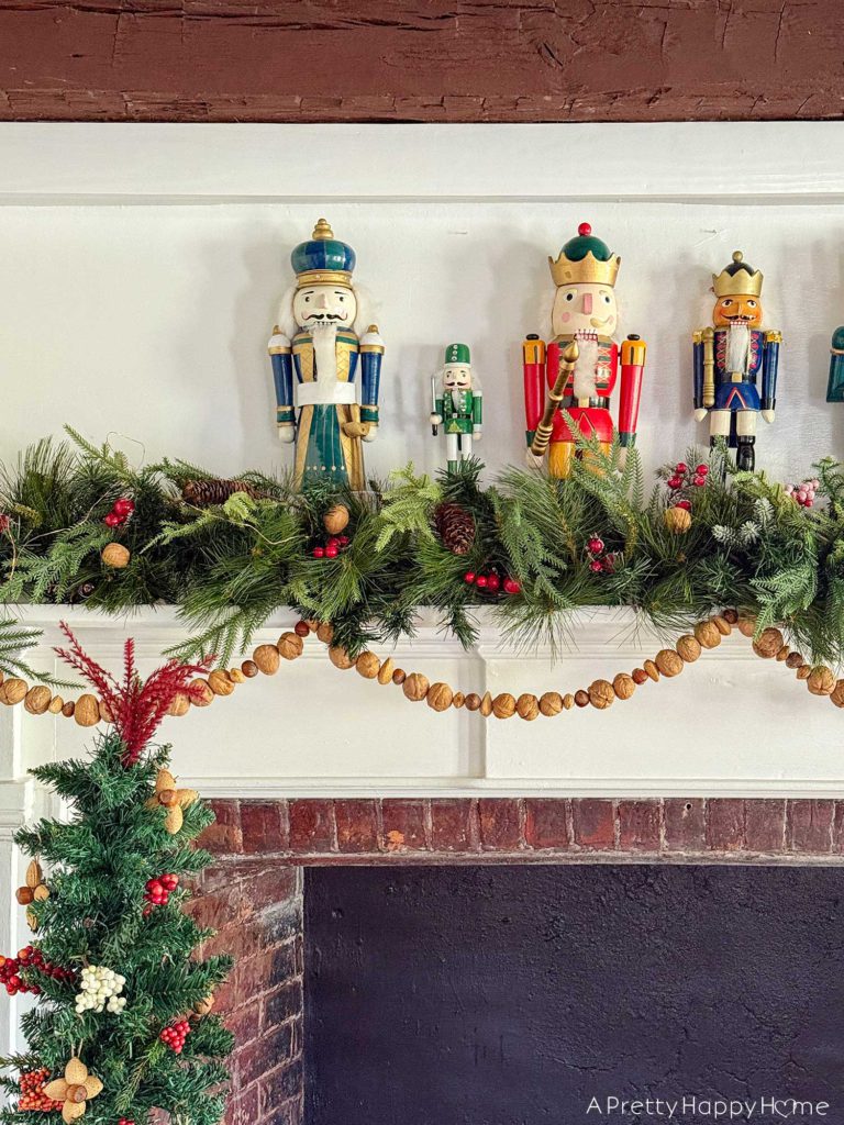 large fireplace in an old house with brick surround decorated for christmas with seven nutcrackers of varying sizes on the mantel and an off centered green garland with red berries. Layered on the mantel is a handmade nut garland comprised of walnuts, hazelnuts, and almonds. Sitting in front of the fireplace is a skinny christmas tree. The tree is decorated with faux red and white berries and ornaments fashioned out of nuts. It is beautiful festive nutcracker christmas mantel decor.