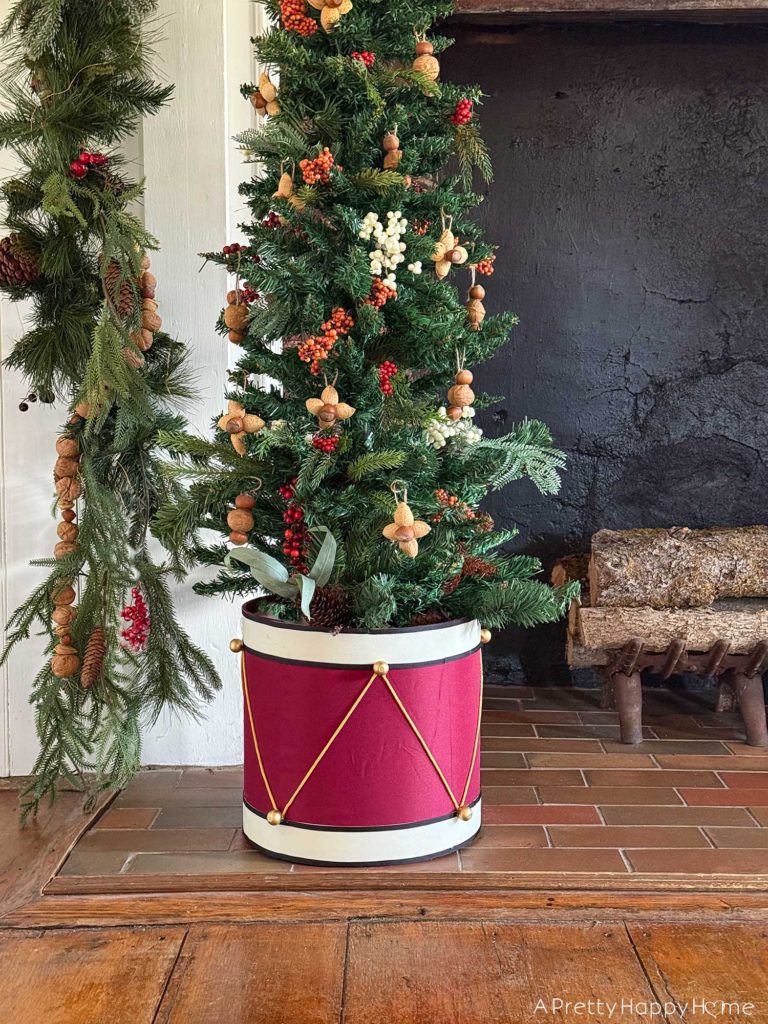 Sitting in front of a fireplace with a brick hearth is a skinny christmas tree with a tree collar that looks like a red drum with gold details. The tree is decorated with faux red and white berries and ornaments fashioned out of nuts. It is beautiful festive nutcracker christmas mantel decor.