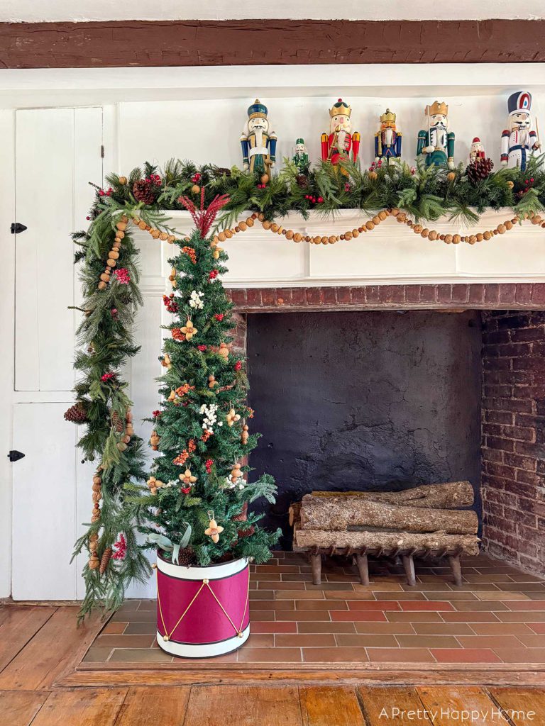 green holiday garland adorning a white fireplace mantel with brick surround. There is a hand strung, in-shell nut garland hanging below the green garland. On top of the mantel are colorful nutcrackers. Sitting in front of the fireplace is a small christmas tree decorated with berries and nut ornaments and it is sitting in a red and white drum. All a part of the blog post five ways to decorate with nuts for christmas.