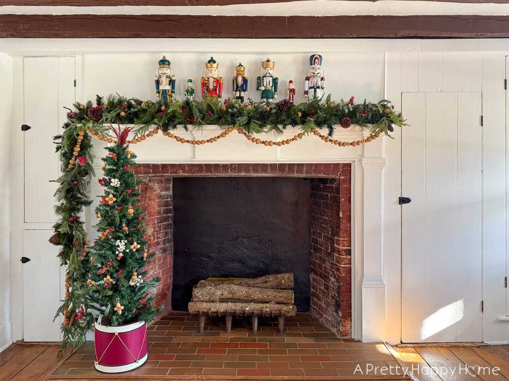 Large fireplace in an old house decorated for christmas with seven nutcrackers of varying sizes on the mantel and an off centered green garland with red berries. Layered on the mantel is a handmade nut garland comprised of walnuts, hazelnuts, and almonds. Sitting in front of the fireplace is a skinny christmas tree with a tree collar that looks like a red drum. The tree is decorated with faux red and white berries and ornaments fashioned out of nuts. It is beautiful festive nutcracker christmas mantel decor.