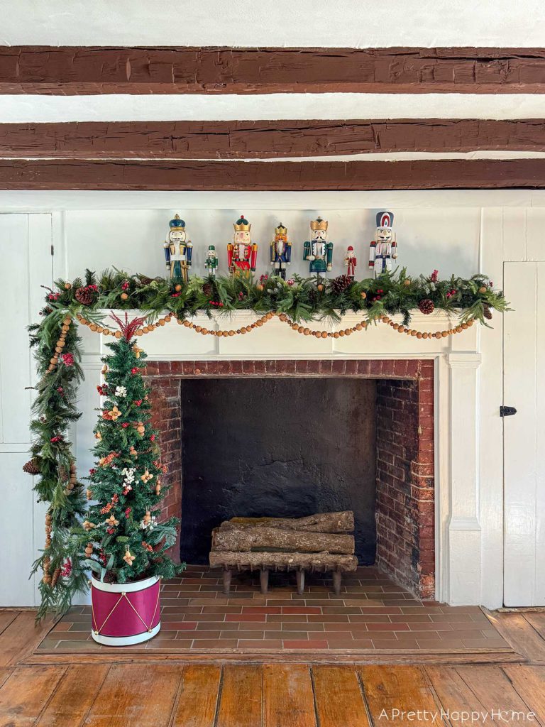 large fireplace in an old house decorated for christmas with seven nutcrackers of varying sizes on the mantel and an off centered green garland with red berries. Layered on the mantel is a handmade nut garland comprised of walnuts, hazelnuts, and almonds. Sitting in front of the fireplace is a skinny christmas tree with a tree collar that looks like a red drum. The tree is decorated with faux red and white berries and ornaments fashioned out of nuts. It is beautiful festive nutcracker christmas mantel decor.