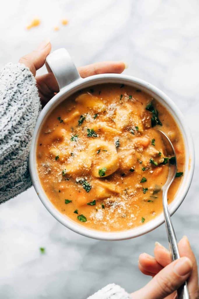 pinch of yum's creamy tortellini soup recipe. image shows a woman's hands holding a bowl of tortellini soup. She's wearing a grey sweater and dipping a spoon into the soup. featured on the happy list