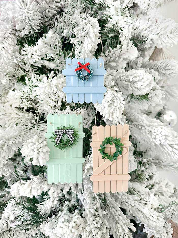 three picket fence ornaments made with popsicle sticks hanging on a flocked christmas tree. The fence ornaments are painted pale blue, mint green, and peach. There are mini wreaths hanging on the picket fence ornaments. Made by Live Laugh Rowe and featured on the happy list