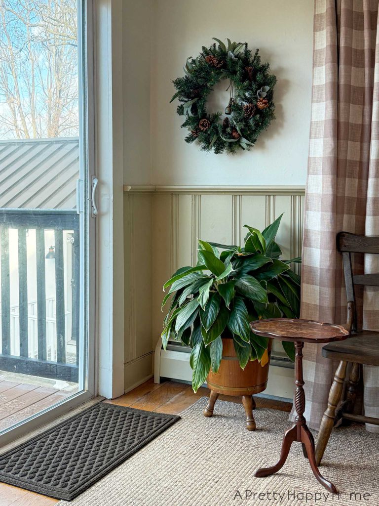 a view of christmas around our home 2025 showing a winter wreath made of fir greenery and pinecones. There are bells hanging in the center of the wreath. The room has a sliding glass door going out to a deck. The wall in the room has a tan color wainscoting, tan buffalo check curtains, a wood drink table, a lush green plant, and a wood bench.