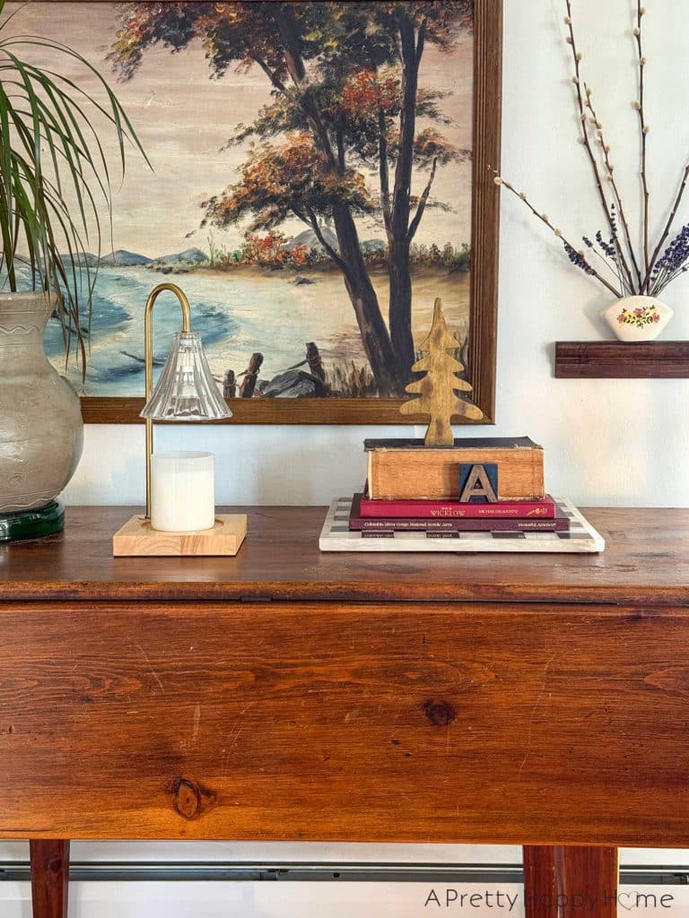 a view of christmas in our home 2025 that shows a living room vignette. There's a drop leaf wood table with a large nature painting of a lake behind the table. Next to the painting is a small wood shelf with a wall pocket. In the wall pocket are dried pussy willows and lavender. On the table is a plant sitting in a dark grey planter. There's a candle warmer lamp with a clear shade. No word on the scent of the candle sitting on the lamp. There's a stack of books on the table with a wood tree sitting on top.