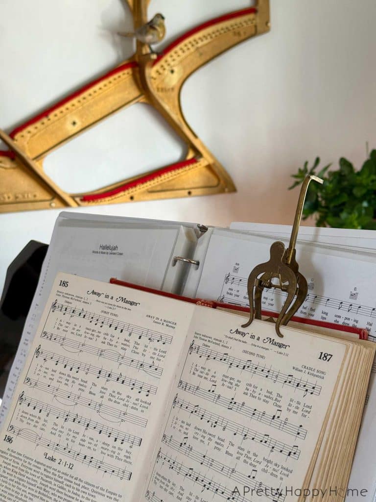 a view of christmas around our home 2025 with a hymnal open on a piano to pages displaying the music to Away in a Manger. IN the background is a brass piano harp hanging on the wall with a faux bird perched on the harp.