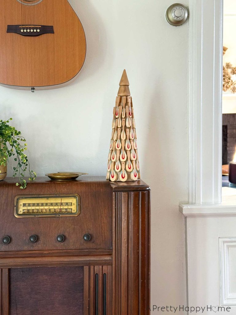 vignette from a music room showing an antique console radio in a dark stain with a small christmas tree made out of repurposed piano hammers sitting on top of the radio. there's a guitar hanging on the wall above the radio. a view of christmas around our home 2025.
