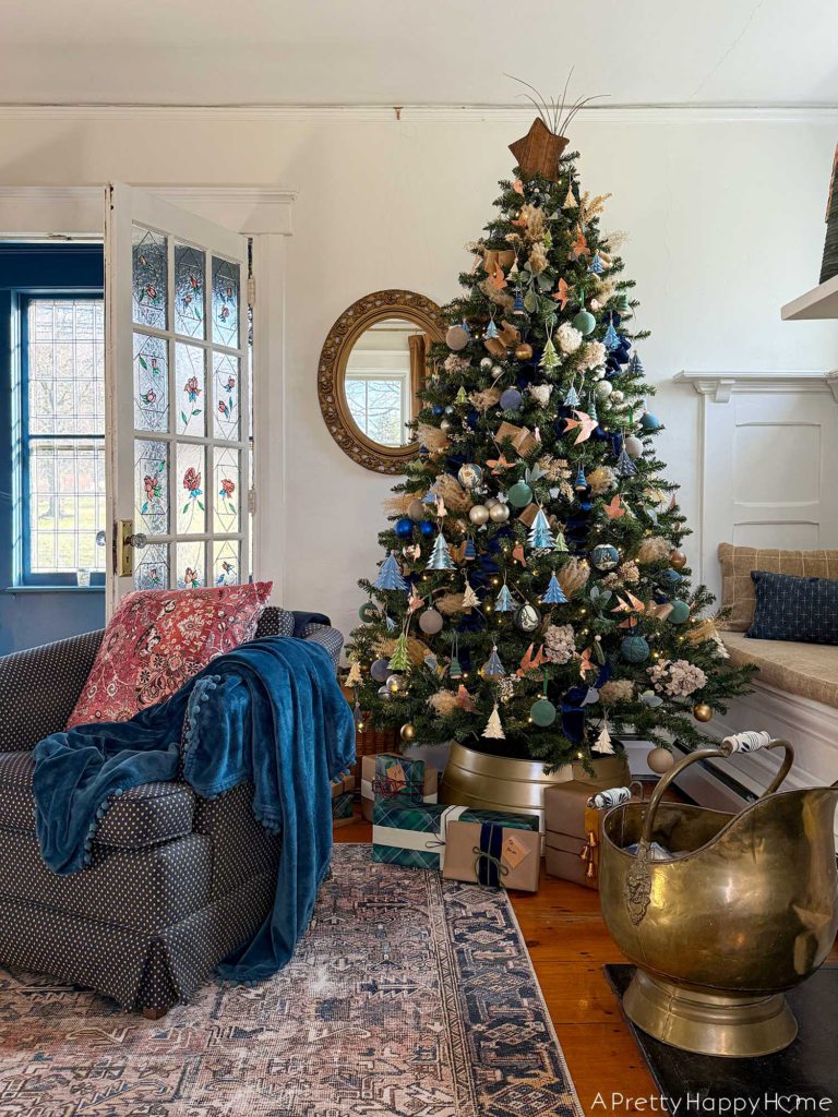 blue and metallic christmas tree decorated with small blue paper tree ornaments, faux velvet baubles in blue and green, small faux velvet tree ornaments in blue and green, pampas grass, blue and tan ribbon, kimekori ornaments, and a wood star topper. The tree is in front of an ornate oval mirror and stained glass french doors.