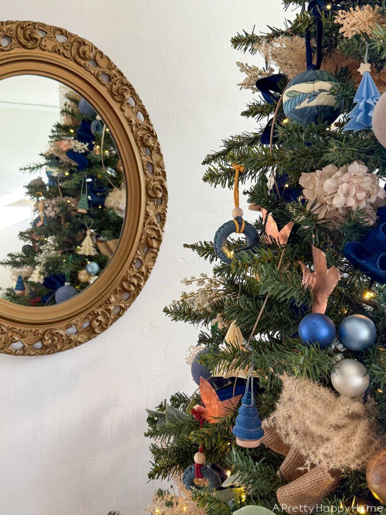 blue and metallic christmas tree decorated with small blue paper tree ornaments, faux velvet baubles in blue and green, small faux velvet tree ornaments in blue and green, pampas grass, blue and tan ribbon, kimekori ornaments, and a wood star topper. The tree is in front of an ornate oval mirror.