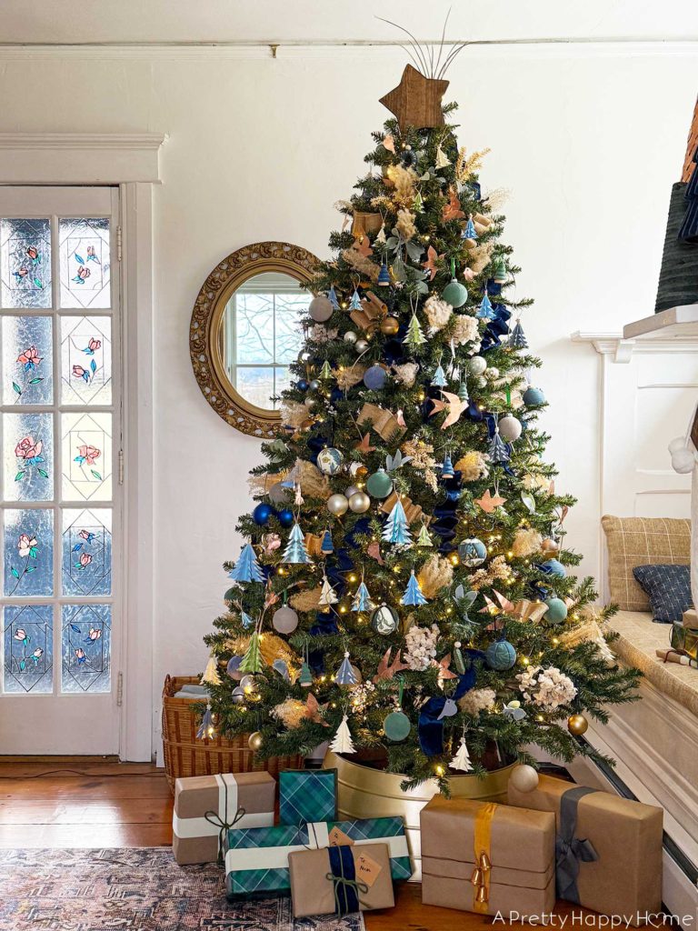 blue and metallic christmas tree decorated with small blue paper tree ornaments, faux velvet baubles in blue and green, small faux velvet tree ornaments in blue and green, pampas grass, blue and tan ribbon, kimekori ornaments, and a wood star topper. The tree is in front of an ornate oval mirror and stained glass french doors.