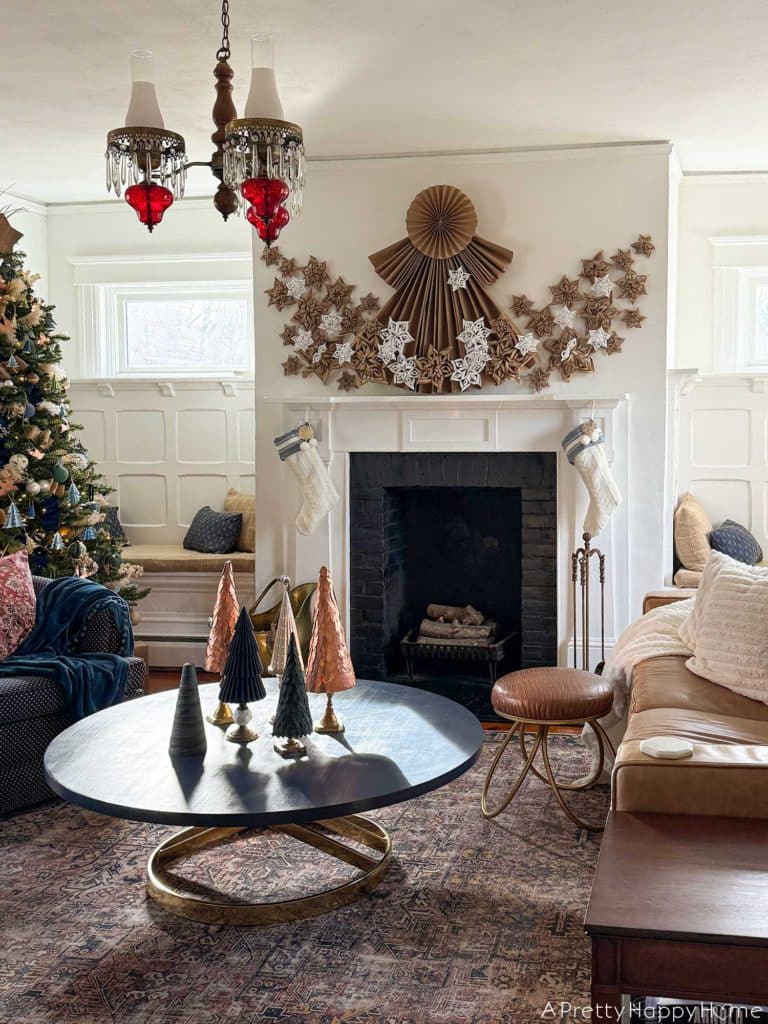 white fireplace with black brick surround decorated for christmas with a 4 foot tall folded giant paper angel made of brown kraft paper. Along the bottom of the angel and fanning out on the sides of her dress are fifty paper snowflakes made from white paper and brown kraft paper. Hanging under the mantel are two white and blue christmas stockings. There's a brass ash bucket in front of the fireplace and a brass fire tool set on the right hand side in front of the fireplace. There's a Christmas tree decorated in blue and green ornaments to the left of the fireplace. In front of the fireplace is a round blue coffee table with six mini Christmas trees sitting on it. Two of the tree are copper, one is white ceramic, one is blue flocked, two are made from green leather.