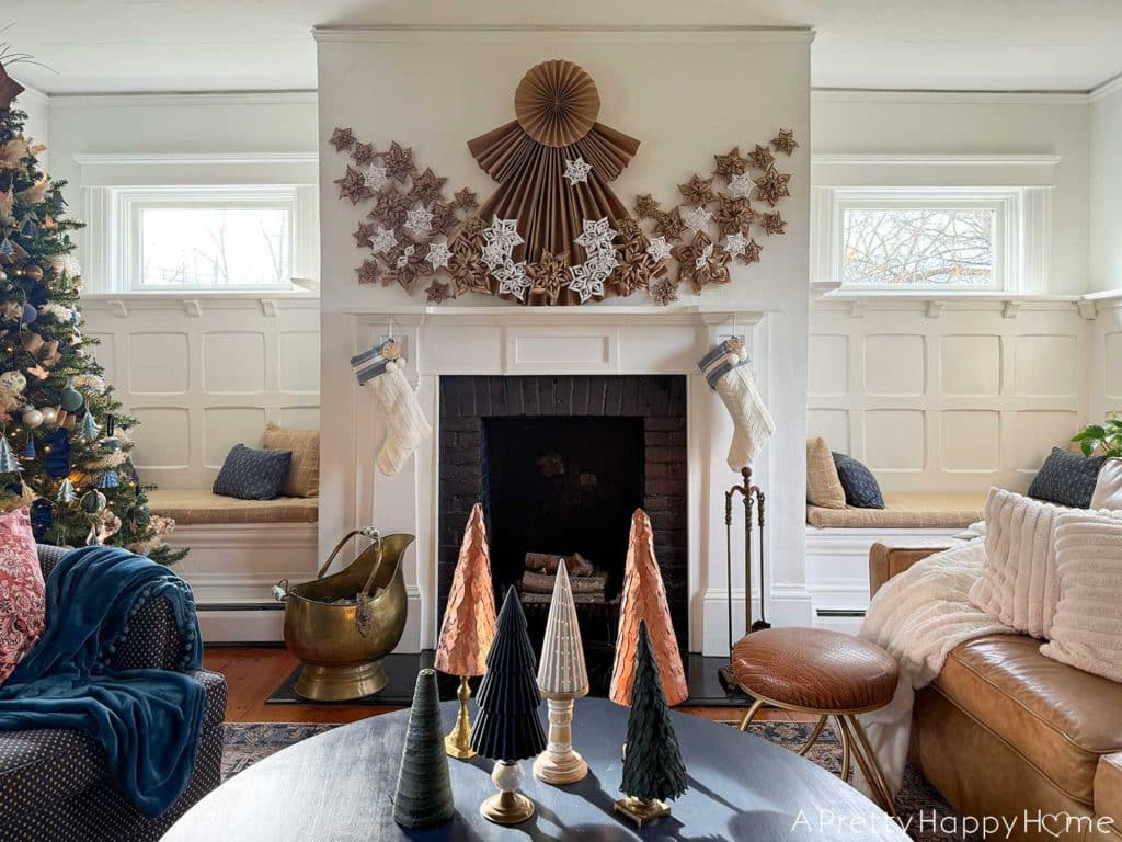 white fireplace with black brick surround decorated for christmas with a 4 foot tall folded giant paper angel made of brown kraft paper. Along the bottom of the angel and fanning out on the sides of her dress are fifty paper snowflakes made from white paper and brown kraft paper. Hanging under the mantel are two white and blue christmas stockings. There's a brass ash bucket in front of the fireplace and a brass fire tool set on the right hand side in front of the fireplace. There's a Christmas tree decorated in blue and green ornaments to the left of the fireplace. In front of the fireplace is a round blue coffee table with six mini Christmas trees sitting on it. Two of the tree are copper, one is white ceramic, one is blue flocked, two are made from green leather.