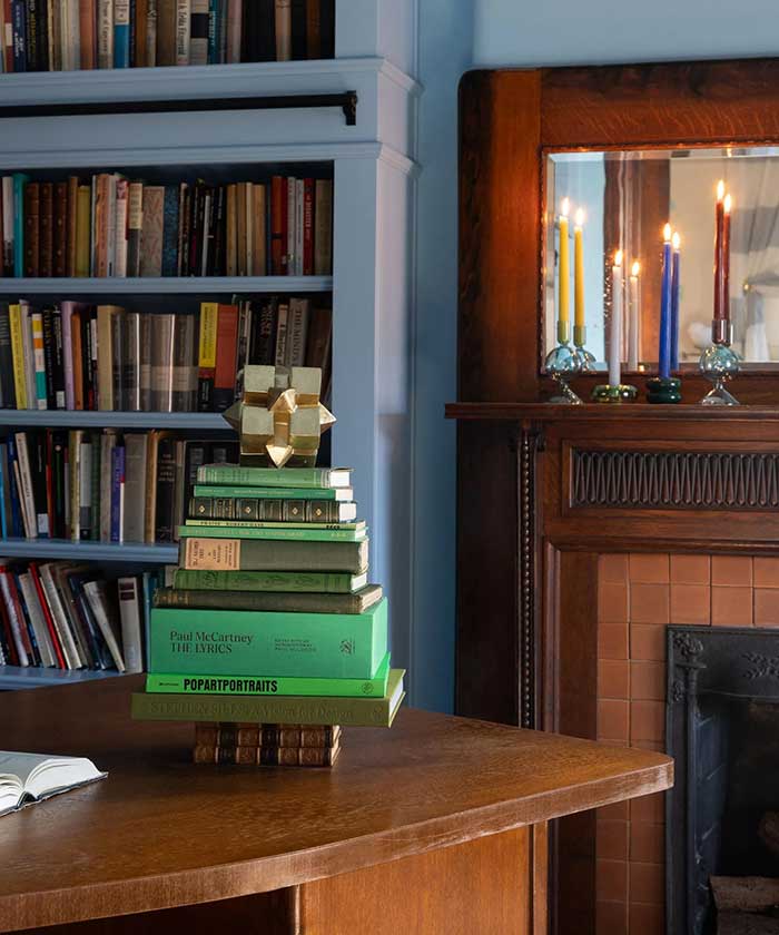 christmas tree made out of a stack of books. Books with a green spine make the body of the tree. Books with a brown spine make the trunk. It sits on a tabletop in front of a built in, light blue bookcase and a fireplace. There are lit candles one red, one blue, one white, on the the fireplace. Image credit to Bethany Adams Interiors and JL Jordan Photography via Homes and Gardens on the happy list.