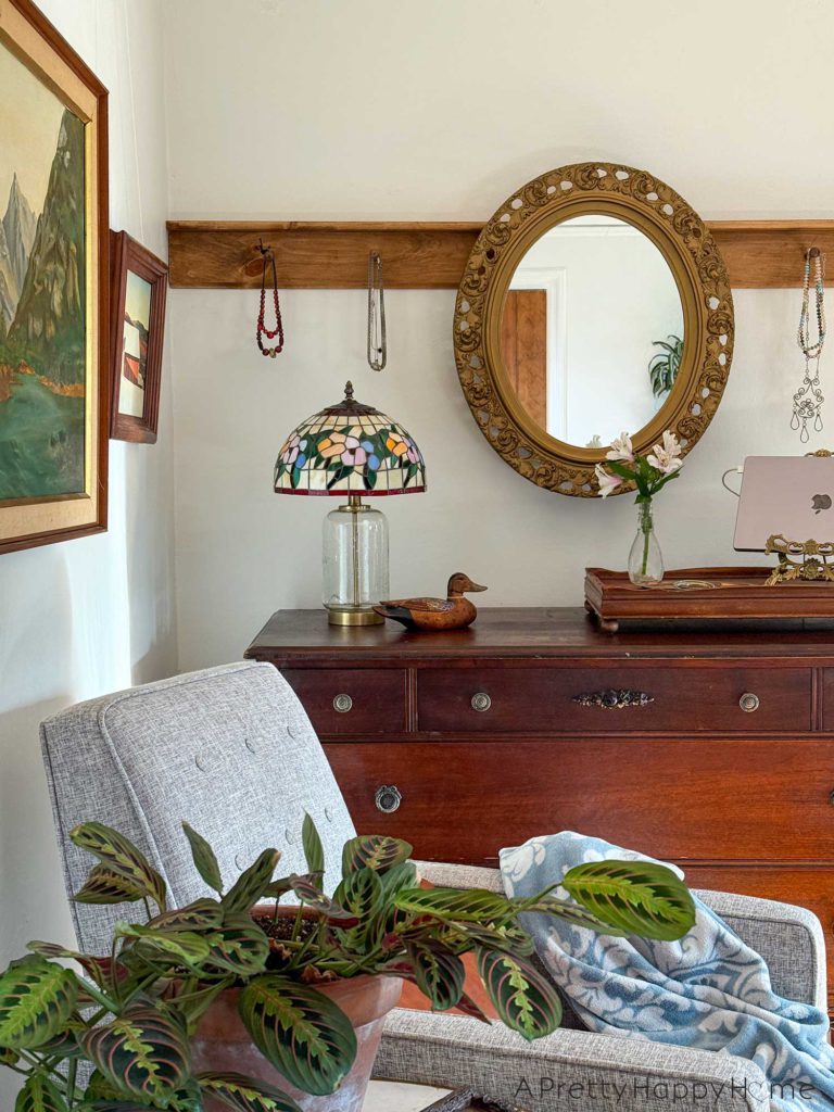 ornate oval mirror hanging from a wood peg rail. There are necklaces on the pegs on either side of the mirror. The mirror hangs above a wood dresser. On the dresser is a tiffany glass lamp, a wood duck, a wood tray with a bouquet of flowers and a laptop stand. In front of the dresser is a seating area with landscape paintings on the wall, grey recliner chairs, a prayer plant, and a light blue throw. The image is showcasing how to hang a mirror from a peg rail.