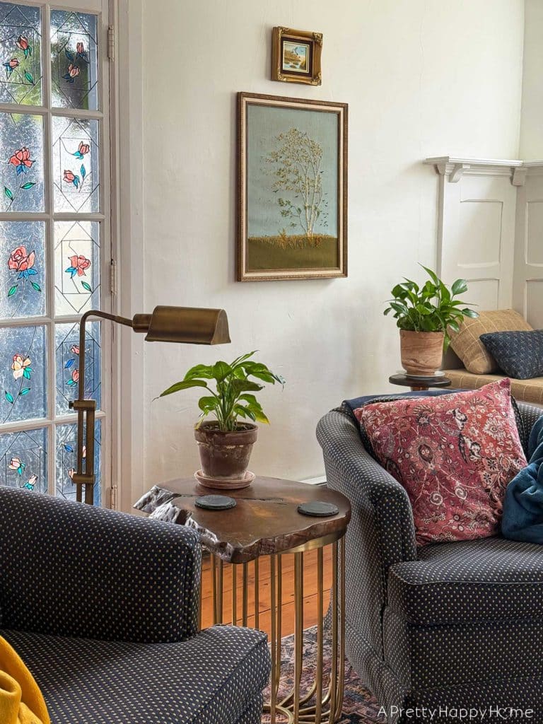 living room vignette focusing on a vintage paragon needlecraft called birch in the wind hanging on the wall. There's a low blue chair sitting in front of the needlepoint. There's also a pothos plant sitting on a wood and brass plant stand in front of the needlepoint, which was thrifted fiber art.