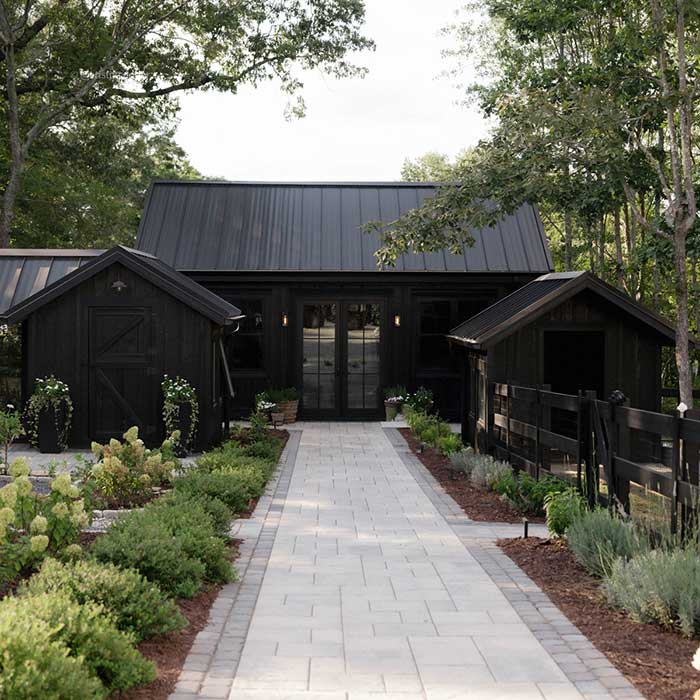cristin cooper's garden with a paver path leading toward a house with black siding. Two small structures also with black siding flank the main house. There are plants growing on the side of the paver path.