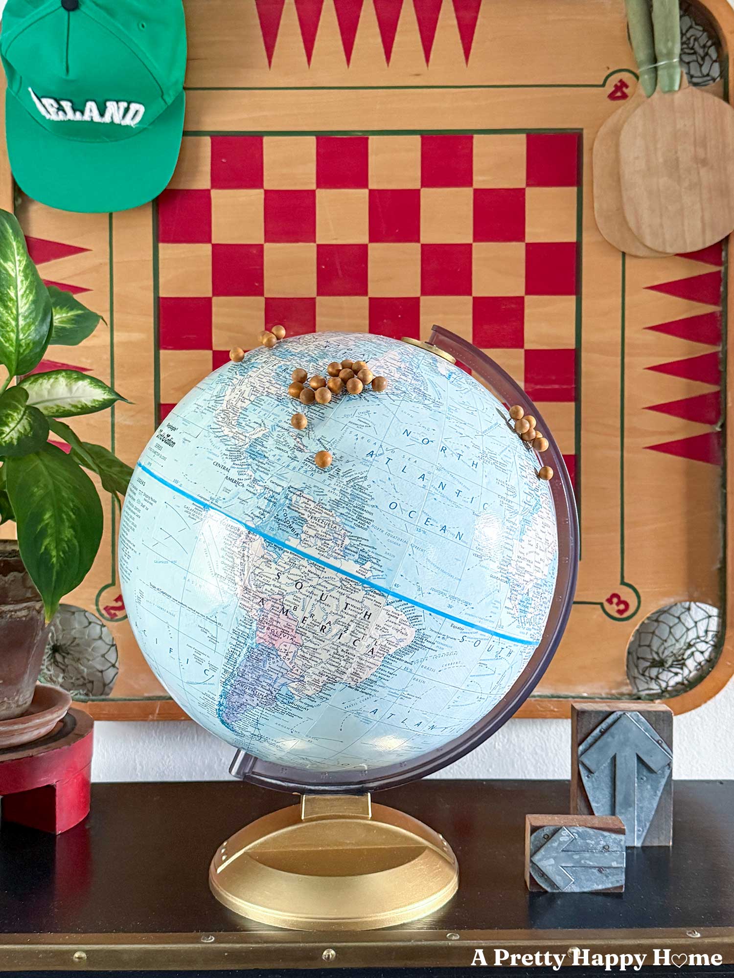 Vintage globe in front of a checkerboard background. The globe has been turned into a diy push pin travel globe with wood push pins inserted into the cardboard globe. There's a green hat of Ireland hanging on the checkerboard with vintage ping pong paddles with green handles hanging next to it.