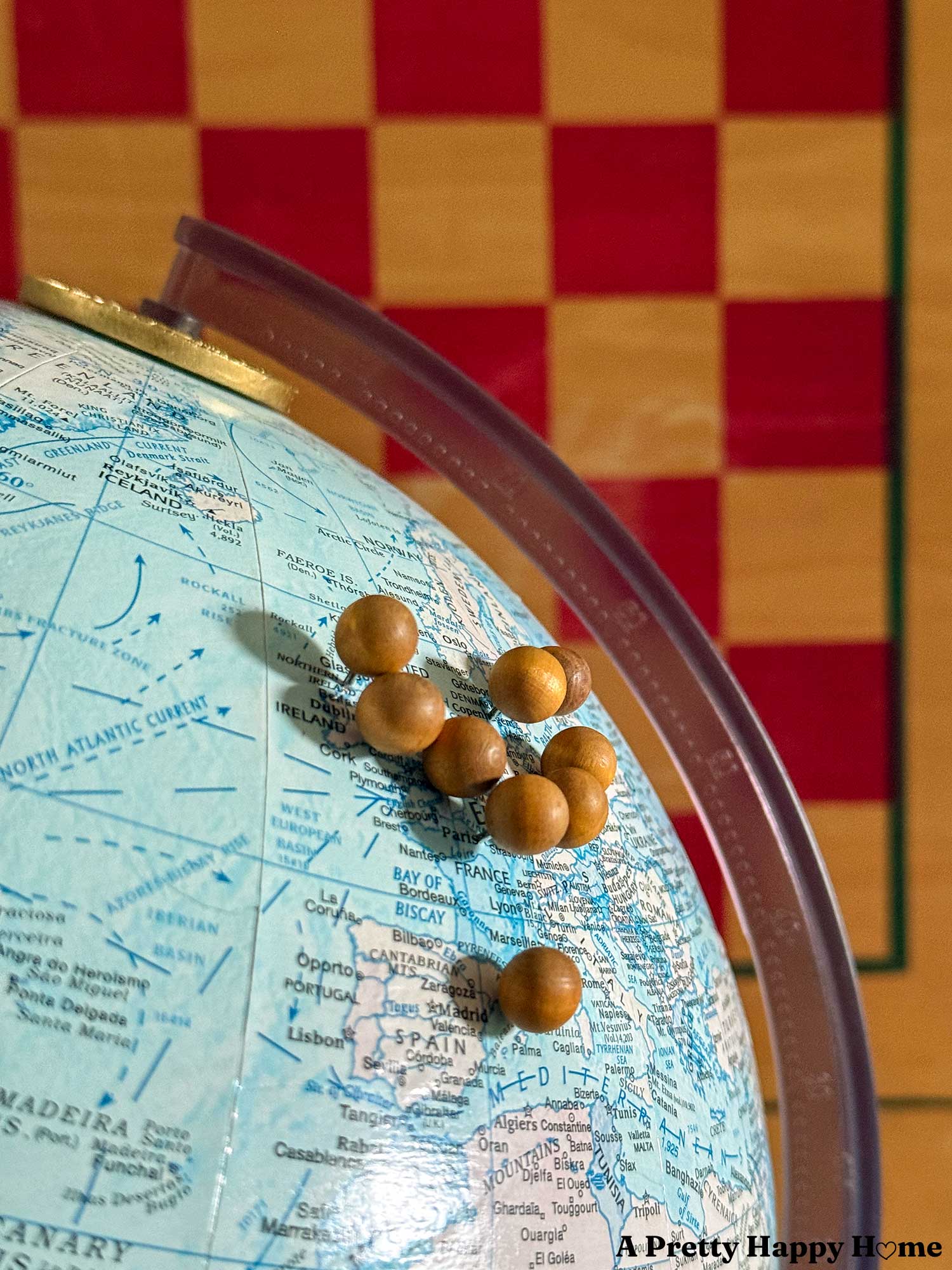 Close up of a vintage globe in front of a checkerboard background. The globe has been turned into a diy push pin travel globe with wood push pins inserted into the cardboard globe.