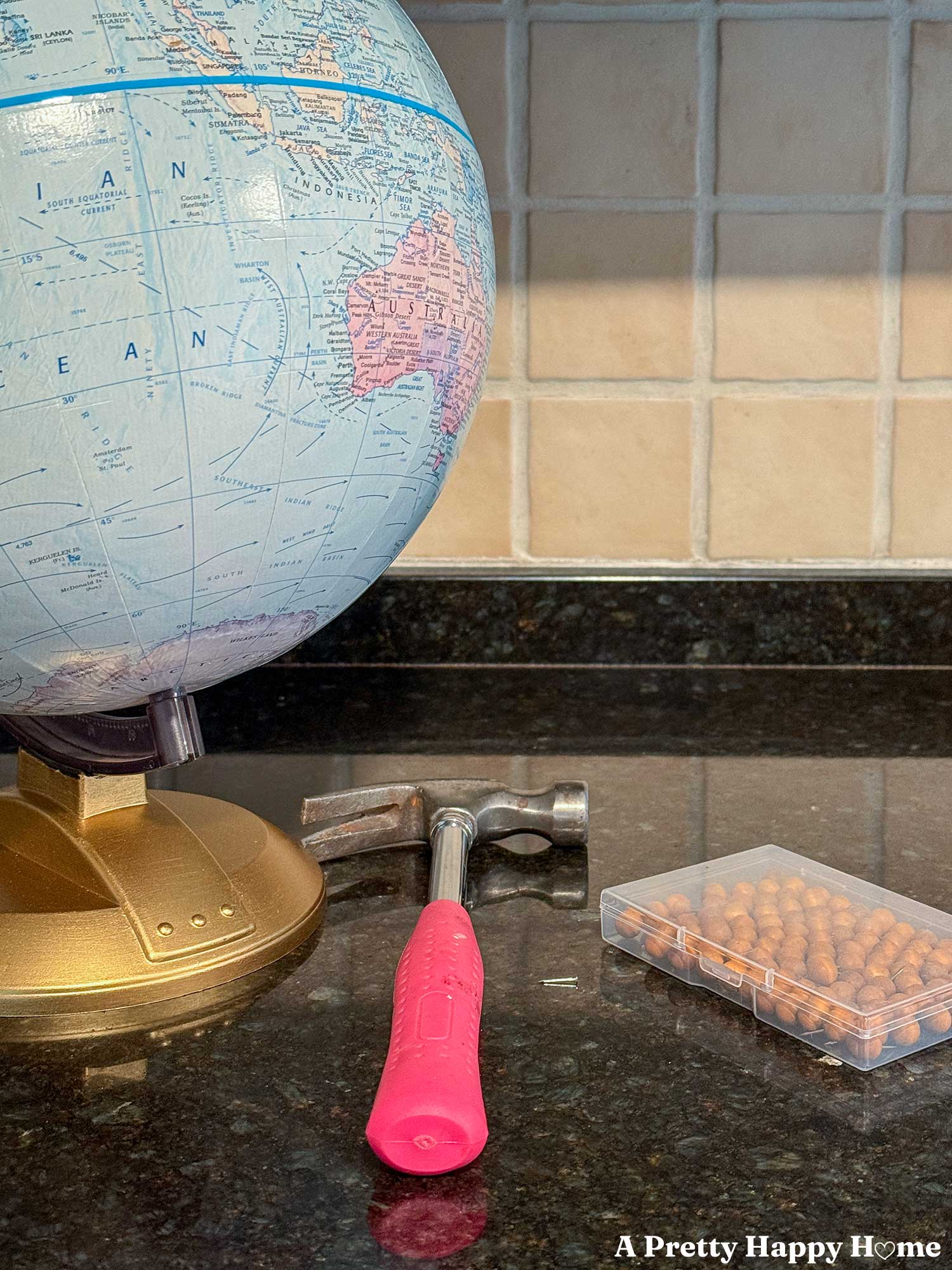 supplies needed to make a diy push pin travel globe including a vintage cardboard globe. This one has a gold base. A hammer. This one has a pink handle. A tiny nail and a packet of push pins or map pins.
