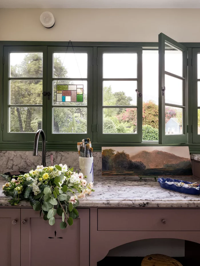 green windows, one of which is open above a marble counter and mauve cabinets with flowers in the sink
